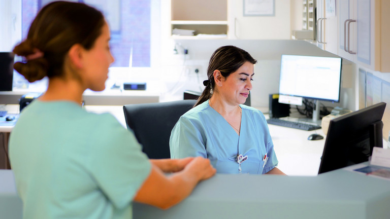 Eine Fachkrankenpflegerin in blauer Funktionskleidung blickt am Empfangstresen des Stationszimmers auf einen Monitor und unterhält sich mit einer anderen Fachkrankenpflegerin im auf Hüftprothesen, Knieprothesen und Schulterprothesen spezialisierten Albertinen Endoprothetikzentrum Hamburg