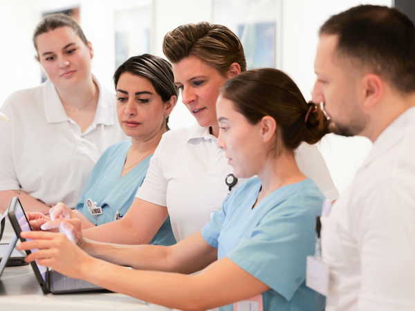 Mehrere Fachkräfte aus dem Team um Zentrumsleiterin Dr. med. Julia M. Reinke blicken auf einen Tablet-Computer auf einem Stationswagen in einem Stationsflur und unterhalten sich im auf Hüftprothesen, Knieprothesen und Schulterprothesen spezialisierten Albertinen Endoprothetikzentrum Hamburg